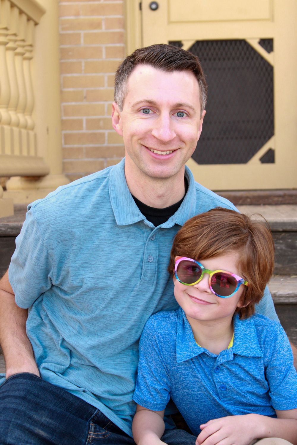 A pair in blue shirts sit together on stone steps near a yellow door.