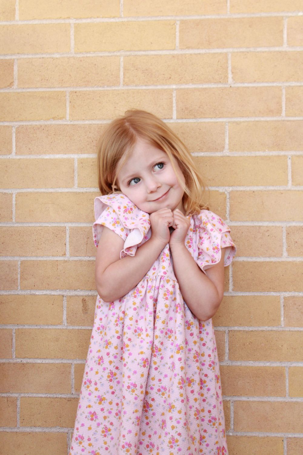 A young child in a pink floral dress poses sweetly against a brick wall.