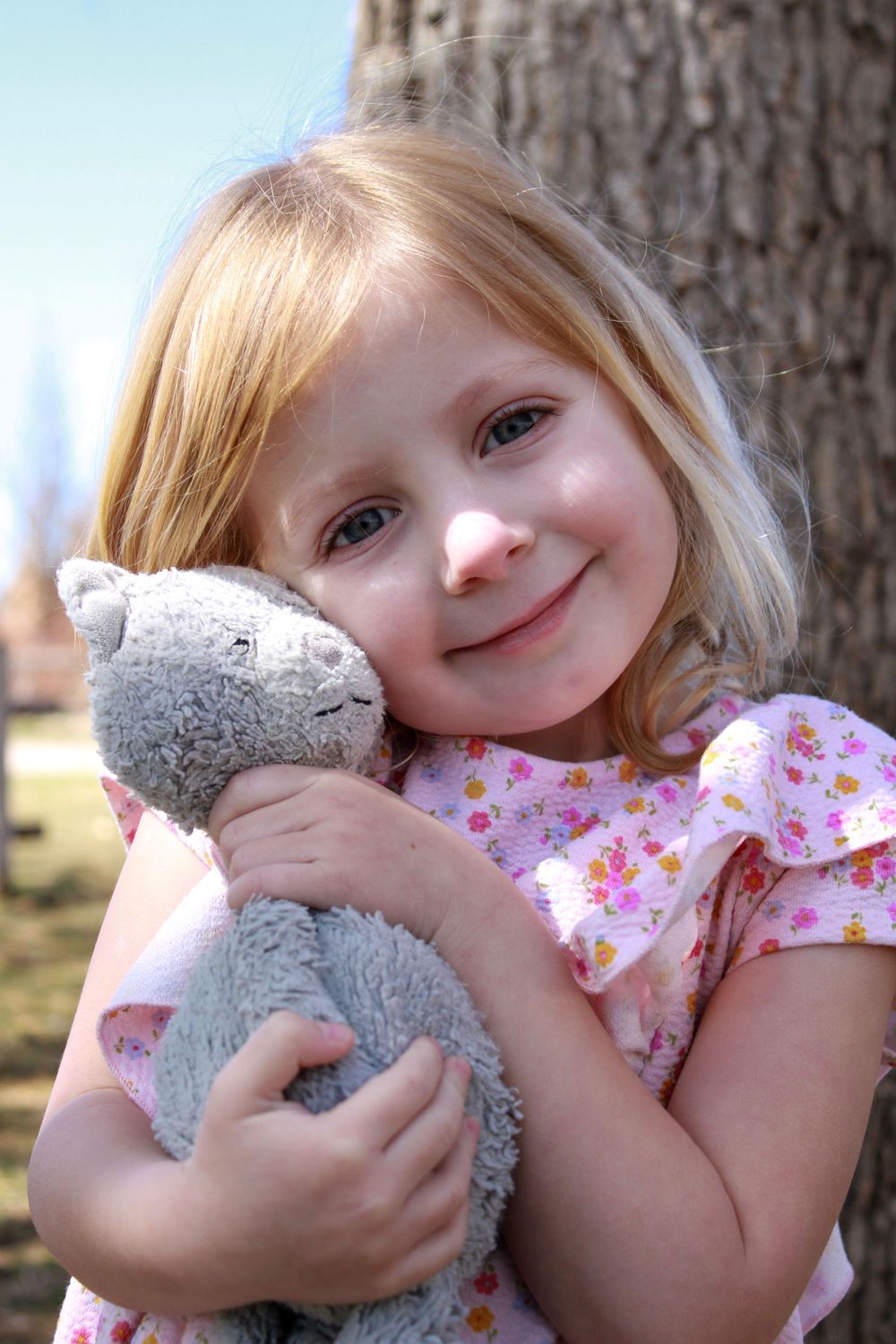 A child in a floral dress hugs a gray stuffed animal while smiling outdoors.