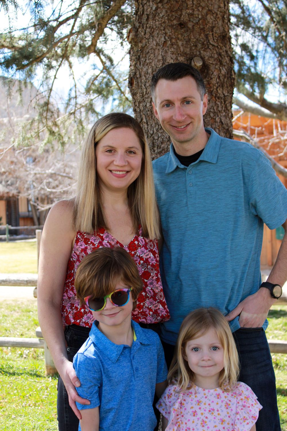 A family of four poses together under a large tree on a sunny day.