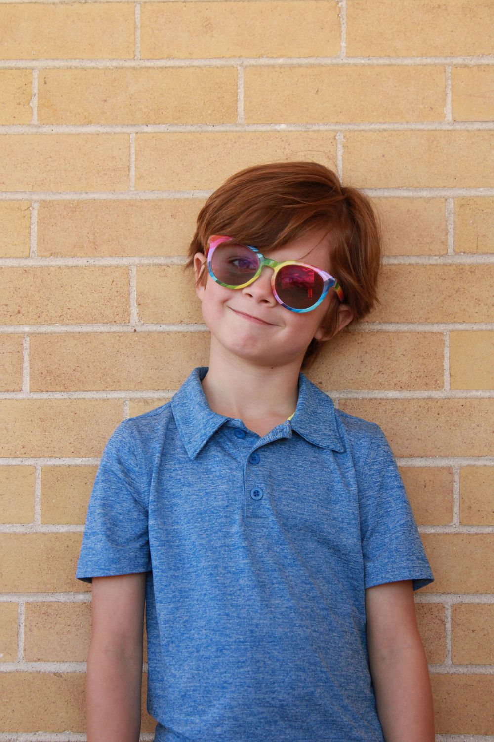 A child wearing colorful sunglasses and blue polo shirt stands against a tan brick wall.