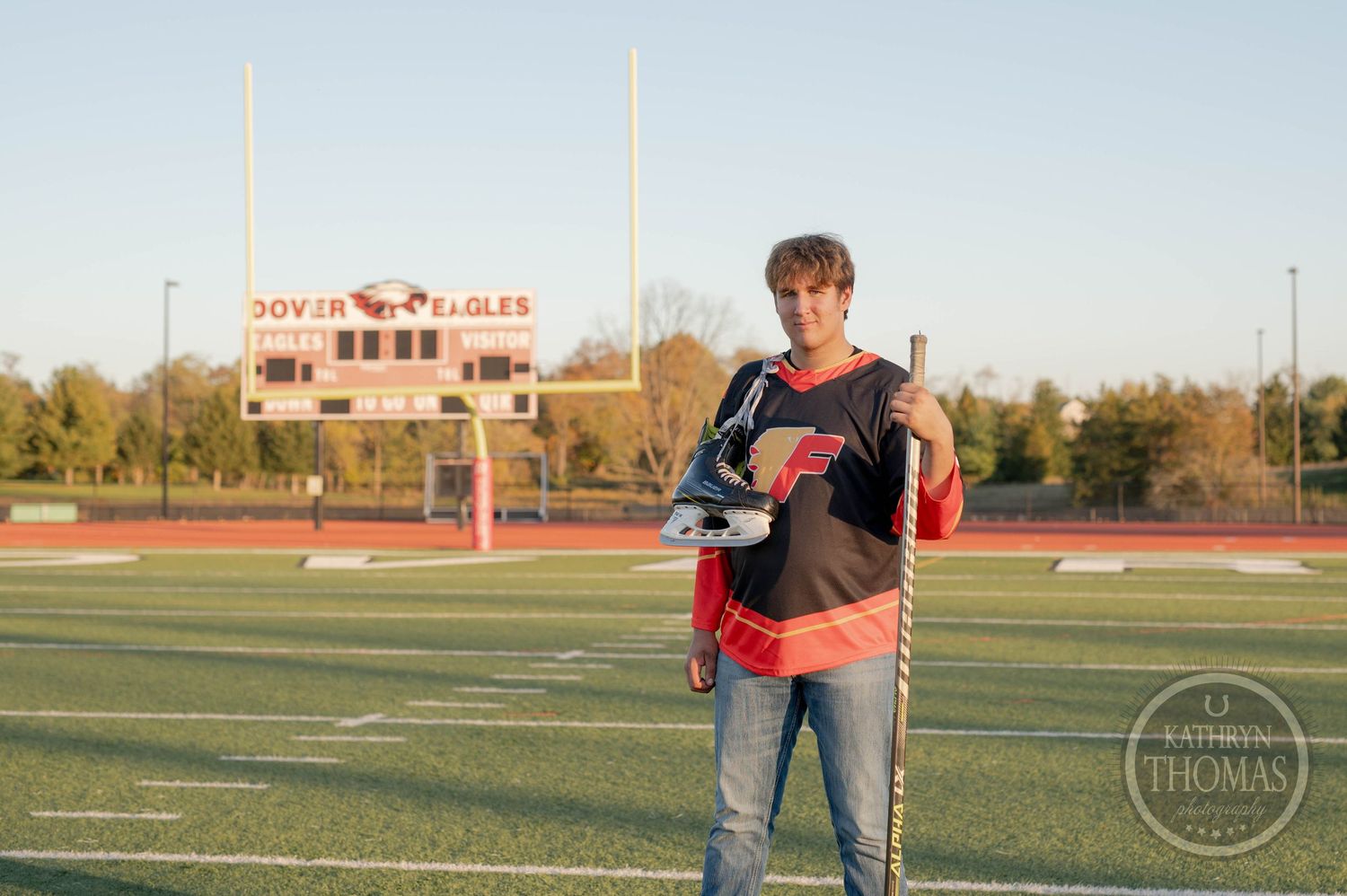 Student athlete poses with hockey gear on football field with stadium scoreboard visible in background.