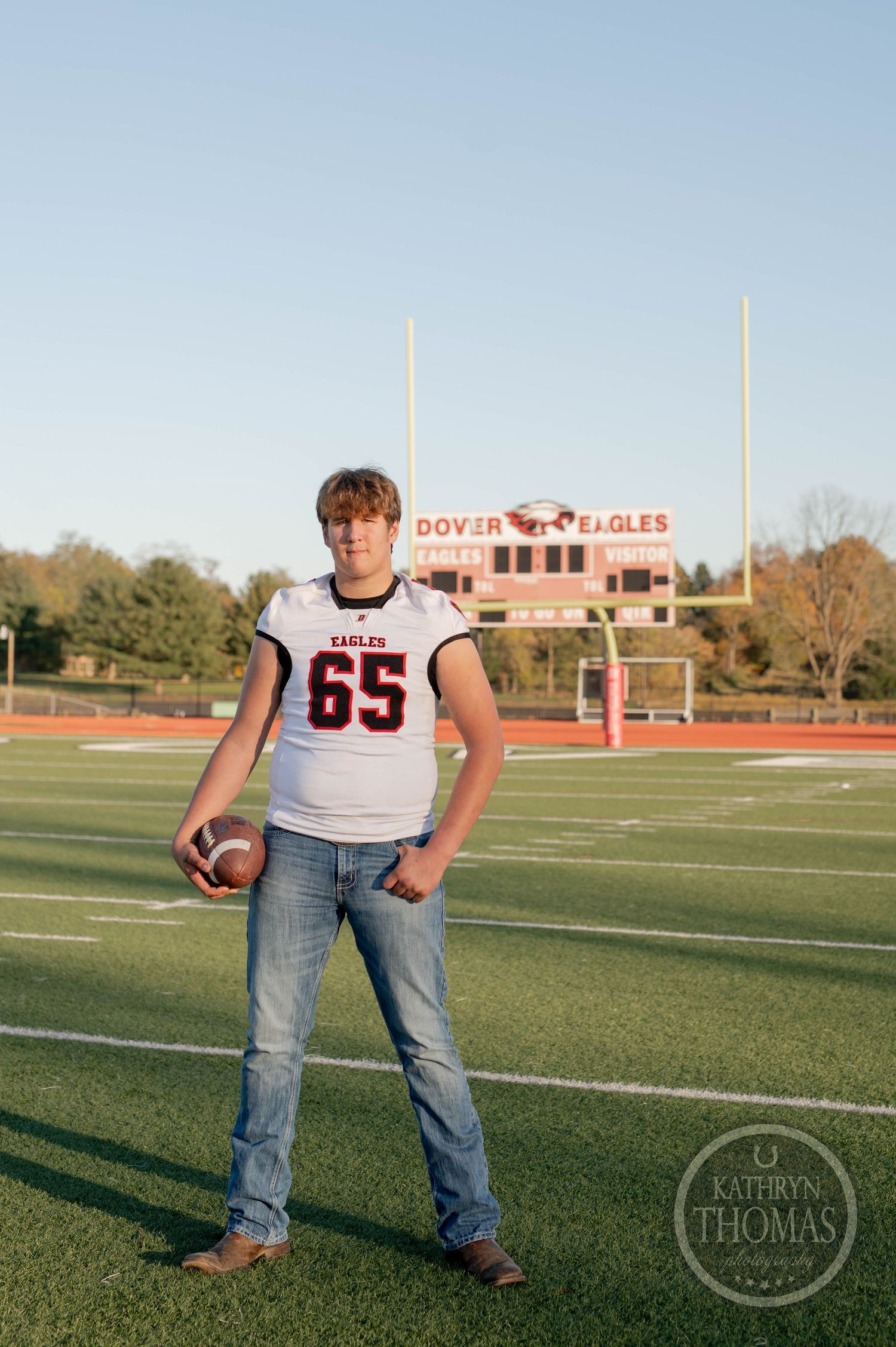 Football player poses with ball on field wearing jersey number 65 with stadium backdrop.