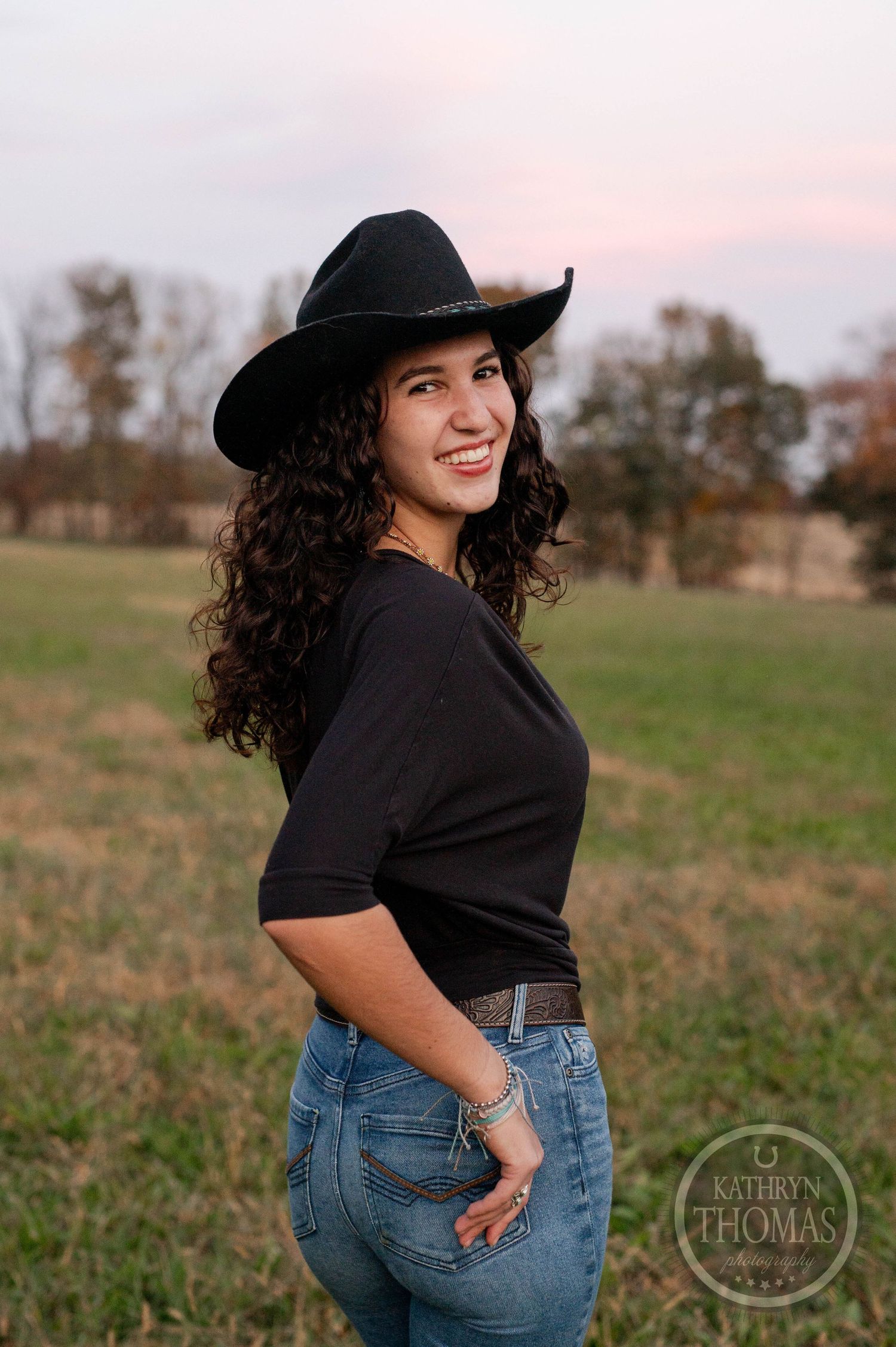 Person in western wear poses against sunset sky in rustic field setting.