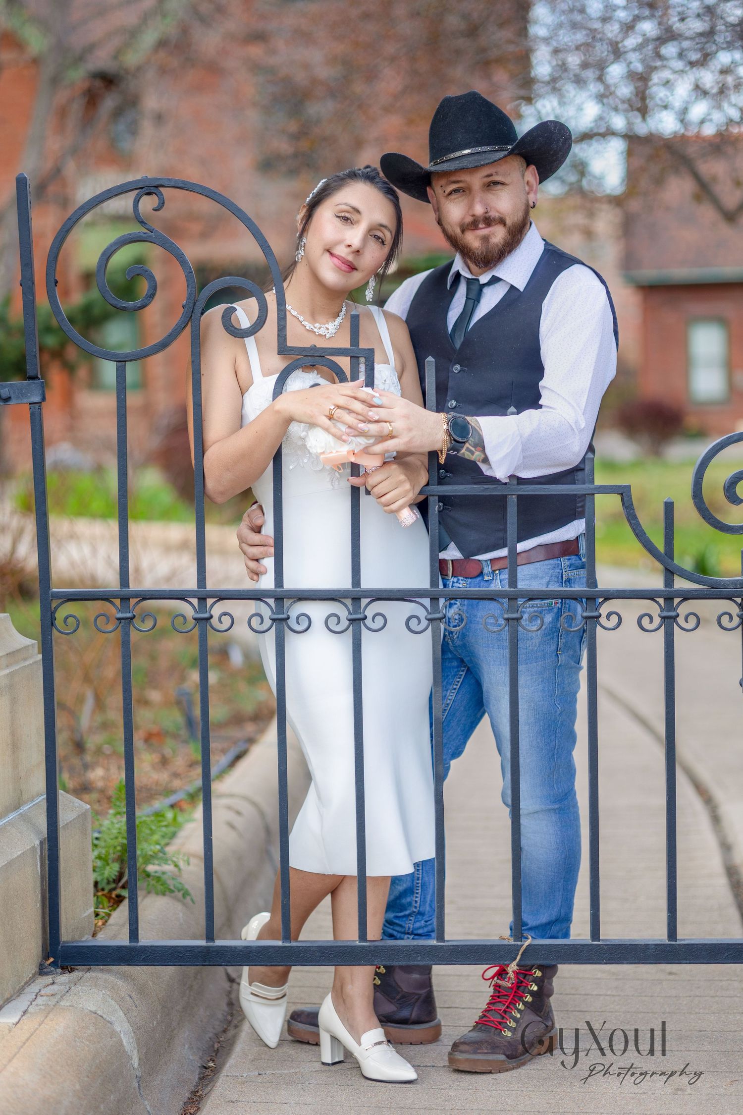 A couple in western attire poses together at an ornate wrought iron gate, dressed in cowboy boots and formal wear.