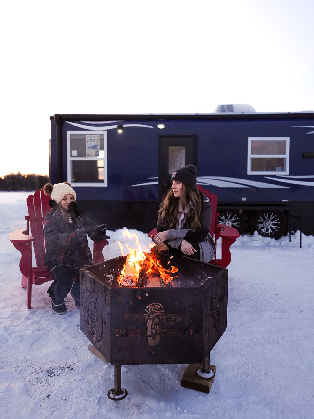 People gathering around a metal fire pit outside a blue ice house in the snow.