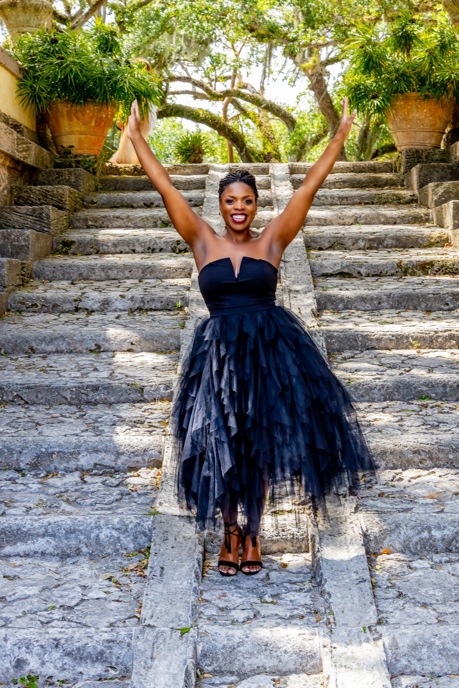 Joyful person in a navy blue tulle dress poses with raised arms on stone steps surrounded by lush greenery.