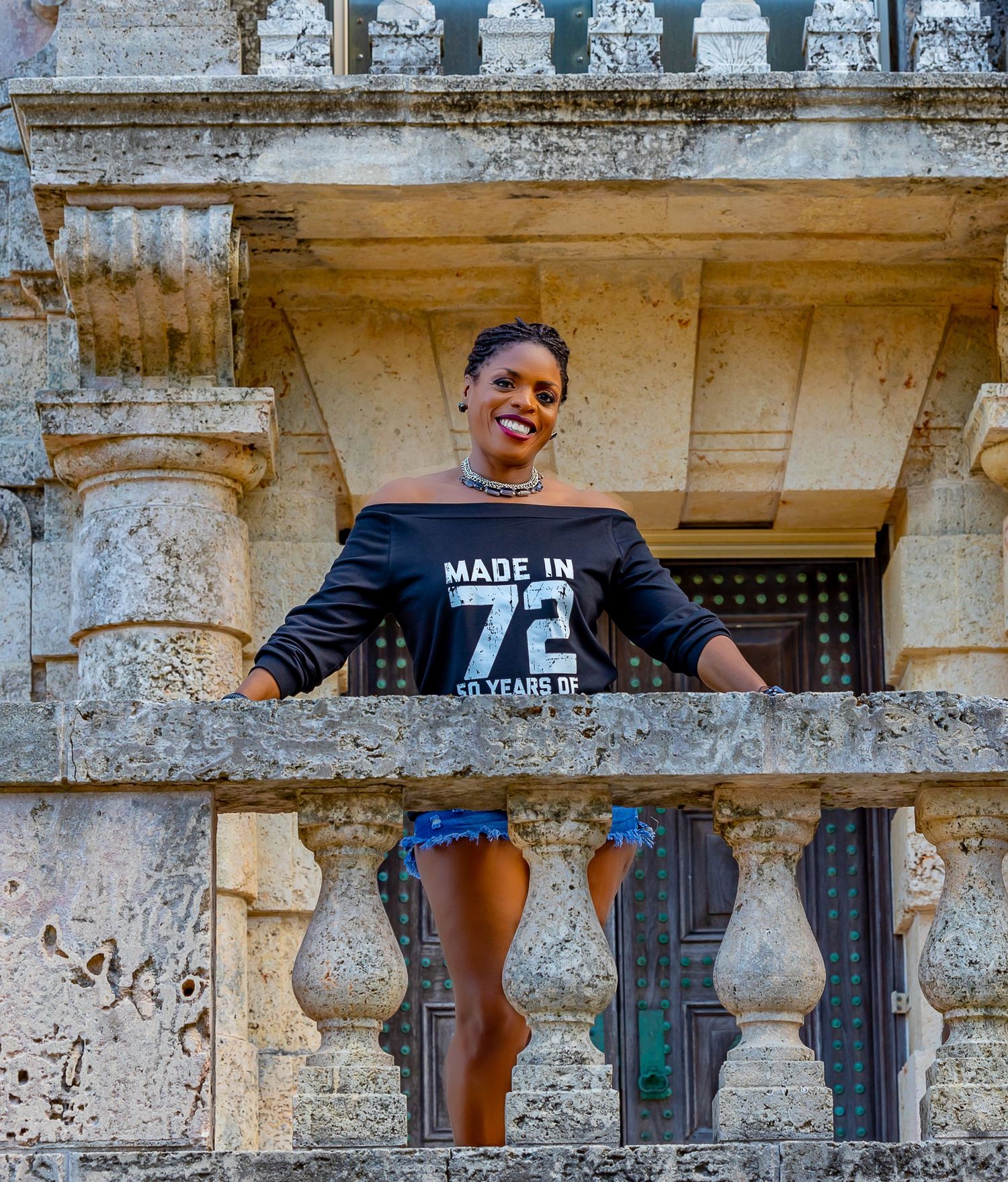Someone wearing a black "Made in 72" shirt stands between ornate stone balustrades of a historic building balcony.
