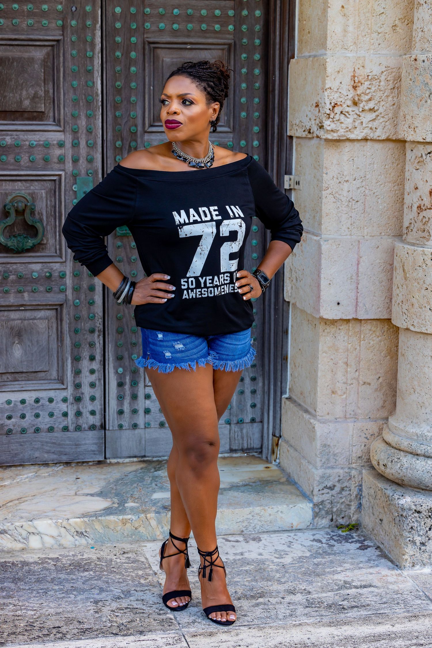 Fashionable off-shoulder black sweater with 'Made in 72' text paired with denim shorts and strappy heels against stone wall.