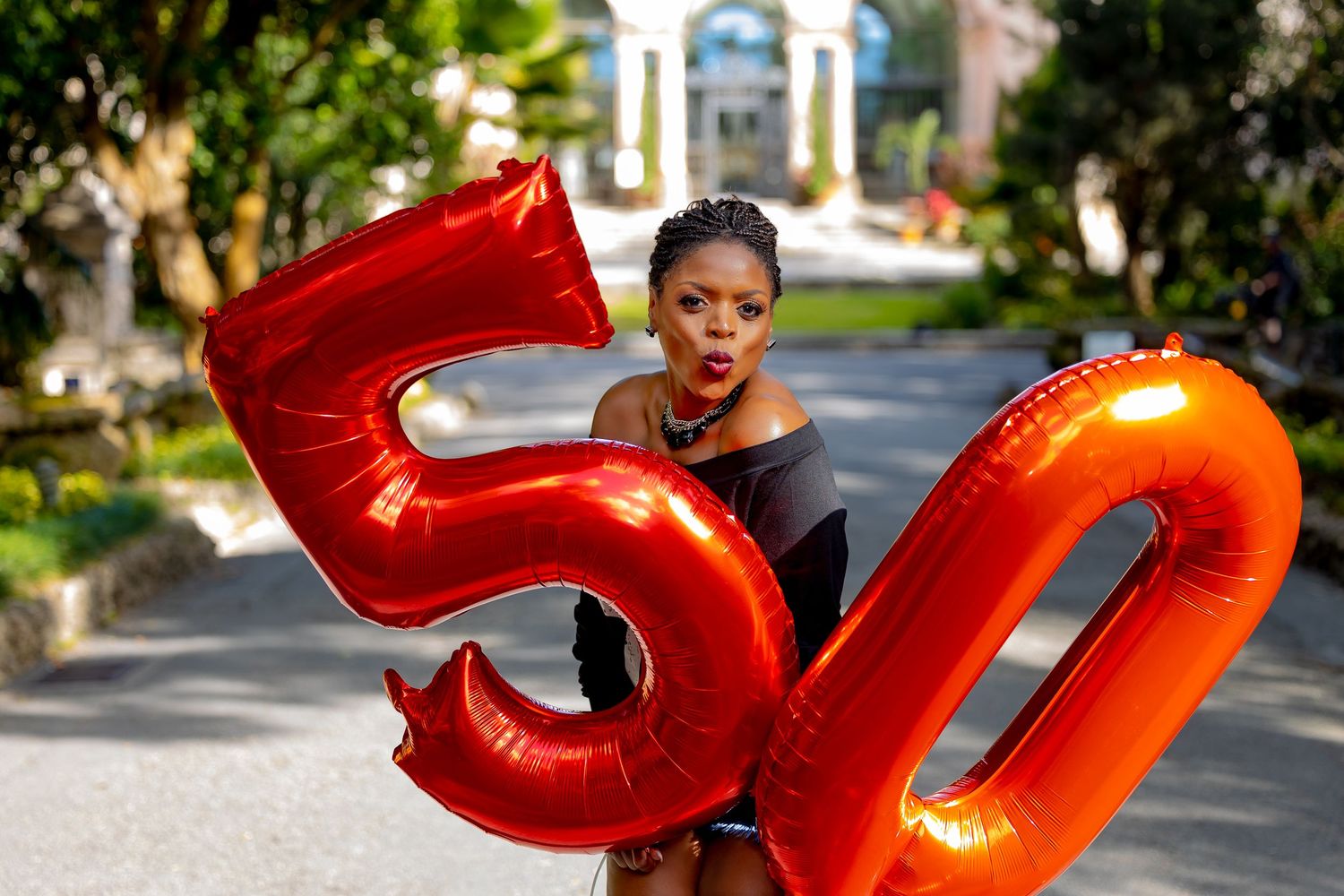 Someone poses with large red metallic number balloons showing 50 in an outdoor setting with greenery and architecture.