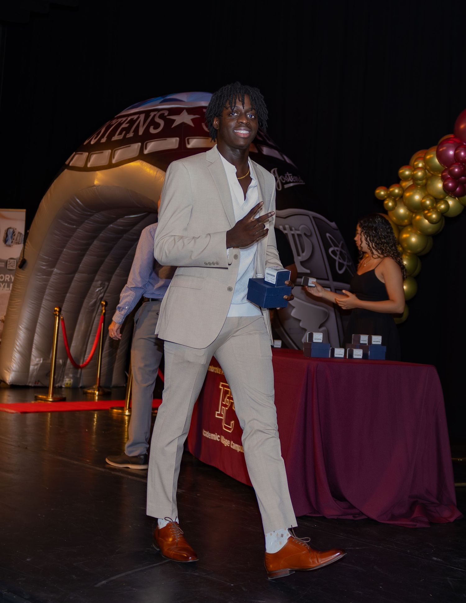 A performer in a white suit dances on stage near inflatable decorations and a burgundy-draped table.