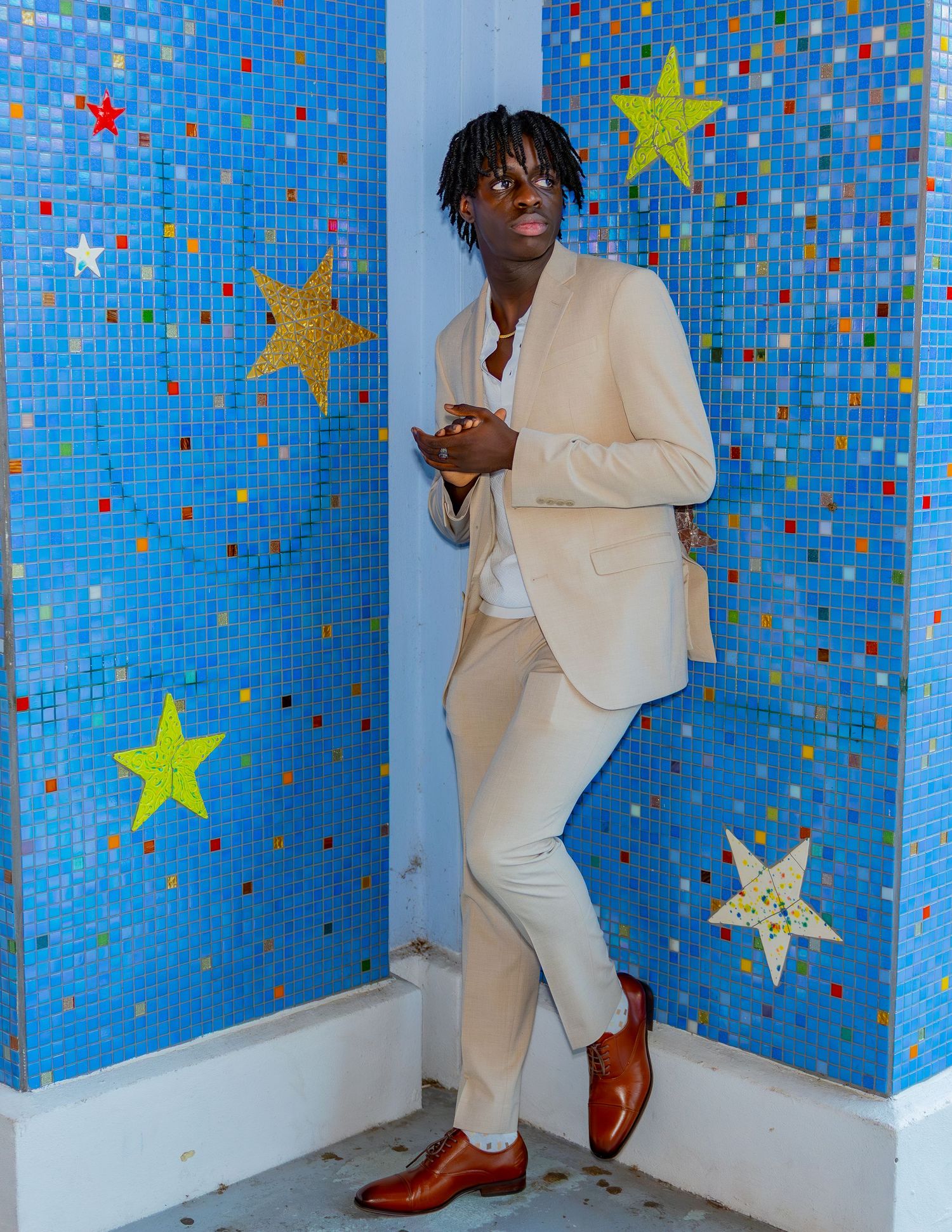 Someone poses against a blue mosaic wall decorated with gold stars while wearing an elegant cream-colored suit and brown shoes.