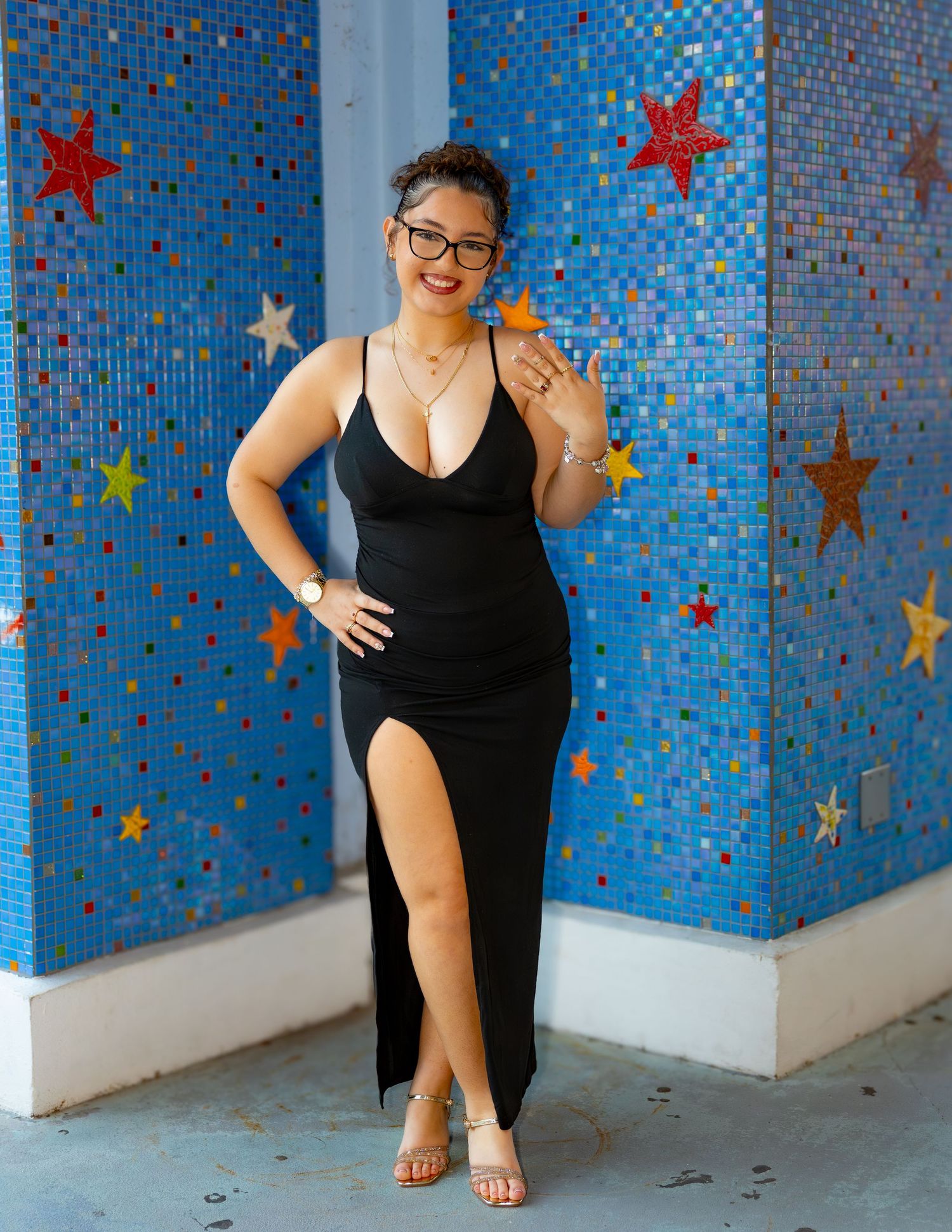 Elegant black evening dress with high slit against blue mosaic wall decorated with colorful stars.