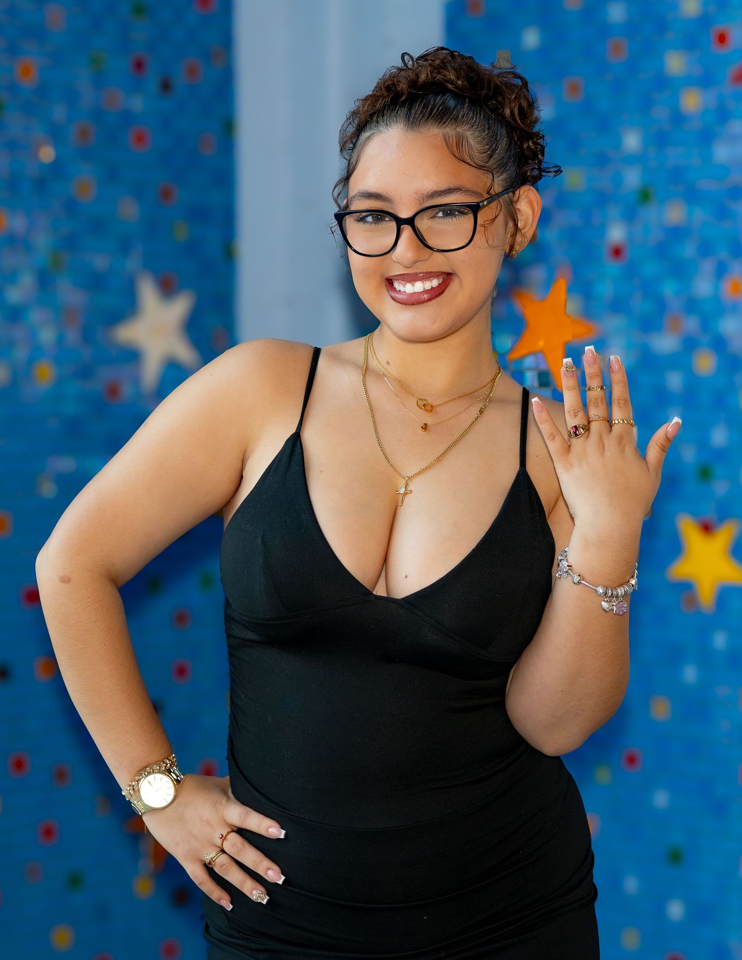 Woman in black sleeveless top poses confidently against blue wall with gold stars, wearing glasses and jewelry.