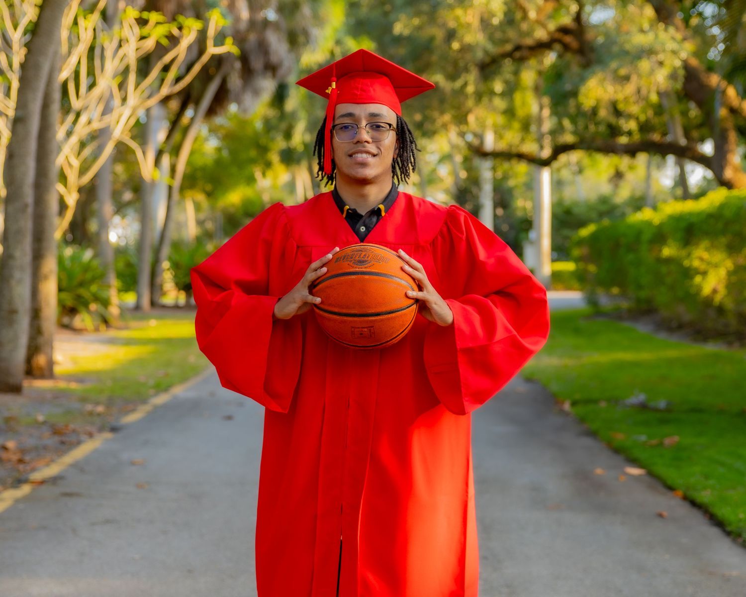 Graduate in bright red cap and gown holds a basketball on tree-lined campus pathway.