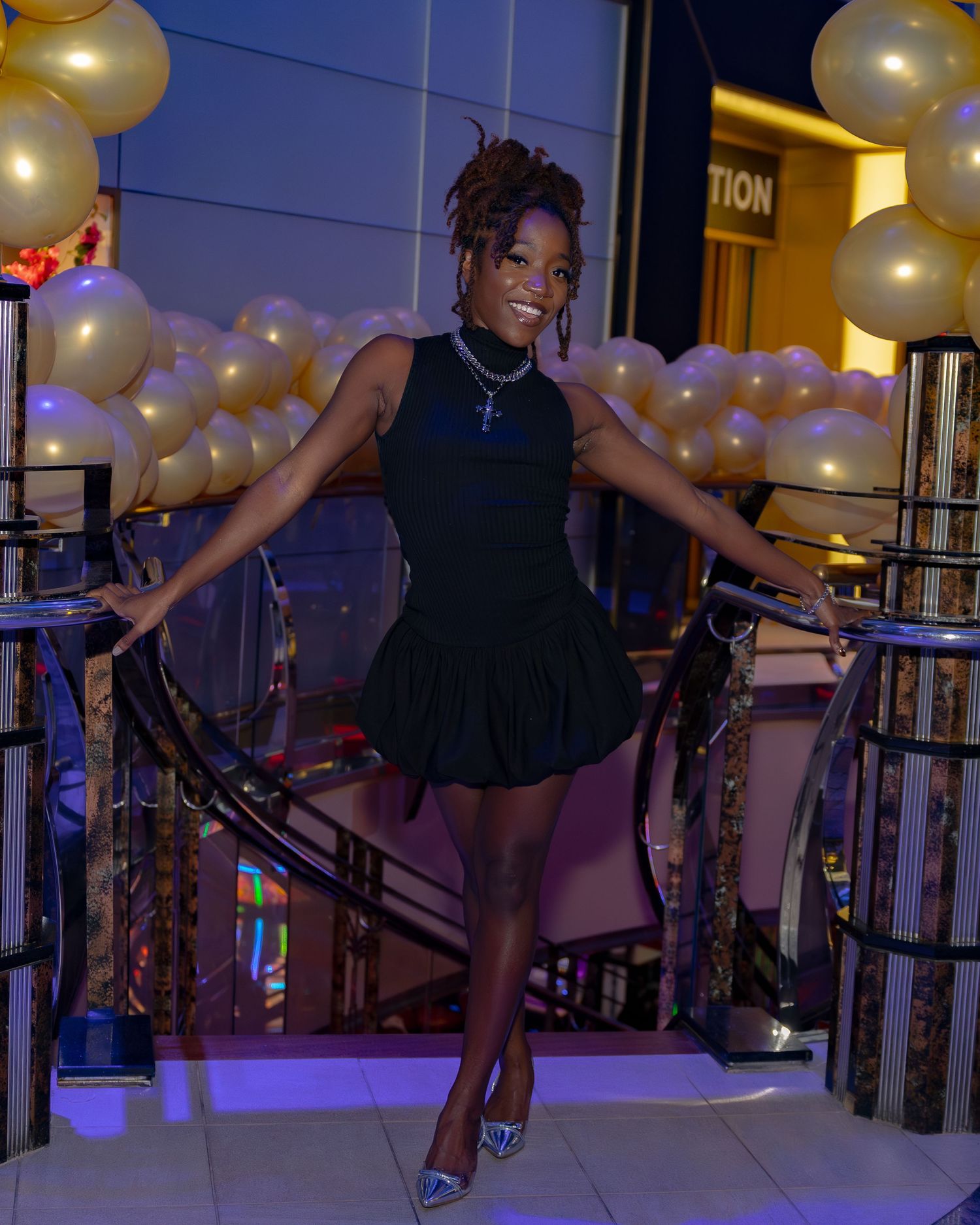 A person in a black sleeveless dress strikes a playful pose by a railing with gold balloons in the background.