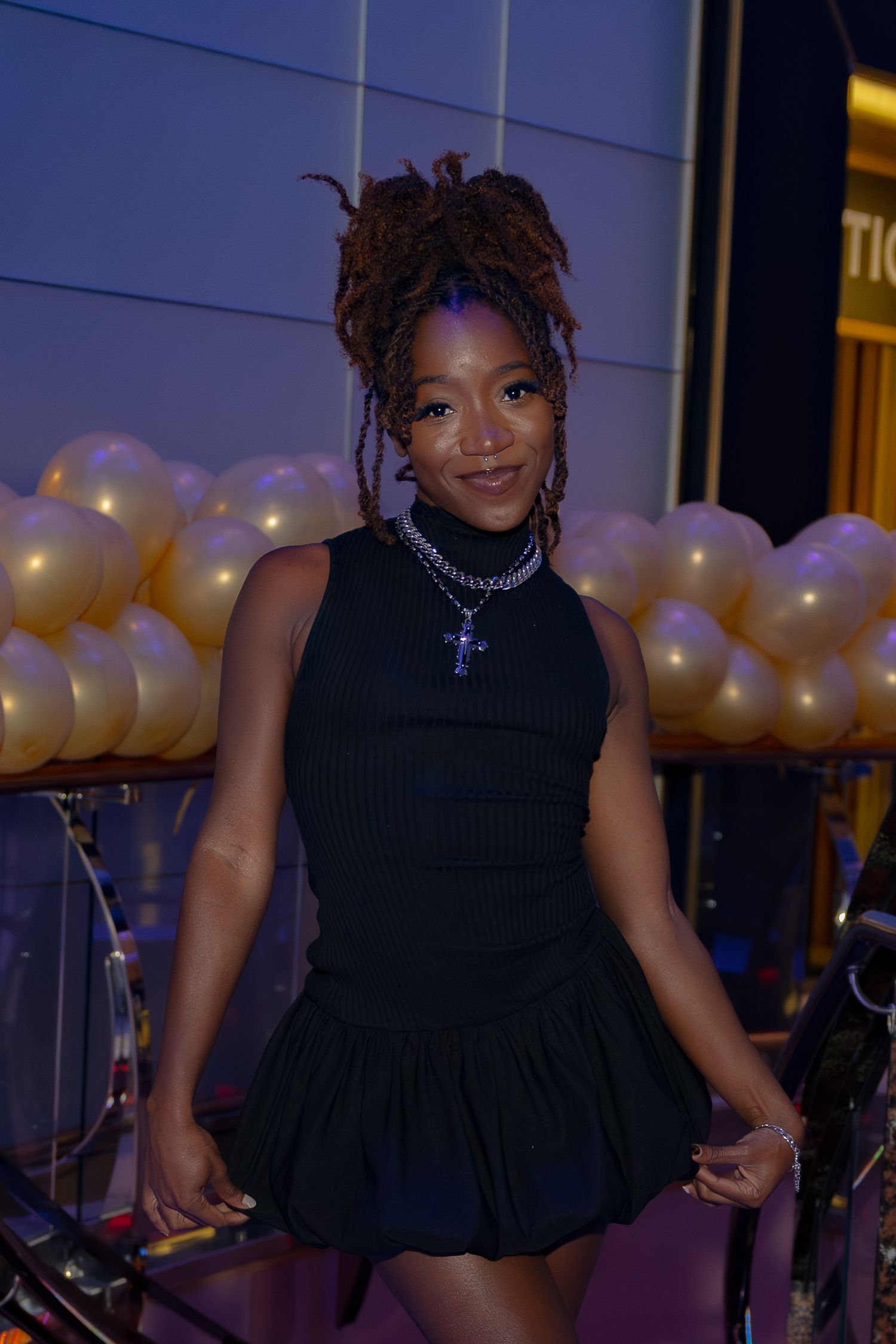 Person in a black sleeveless dress and chunky necklace poses in front of gold balloons at an evening event.