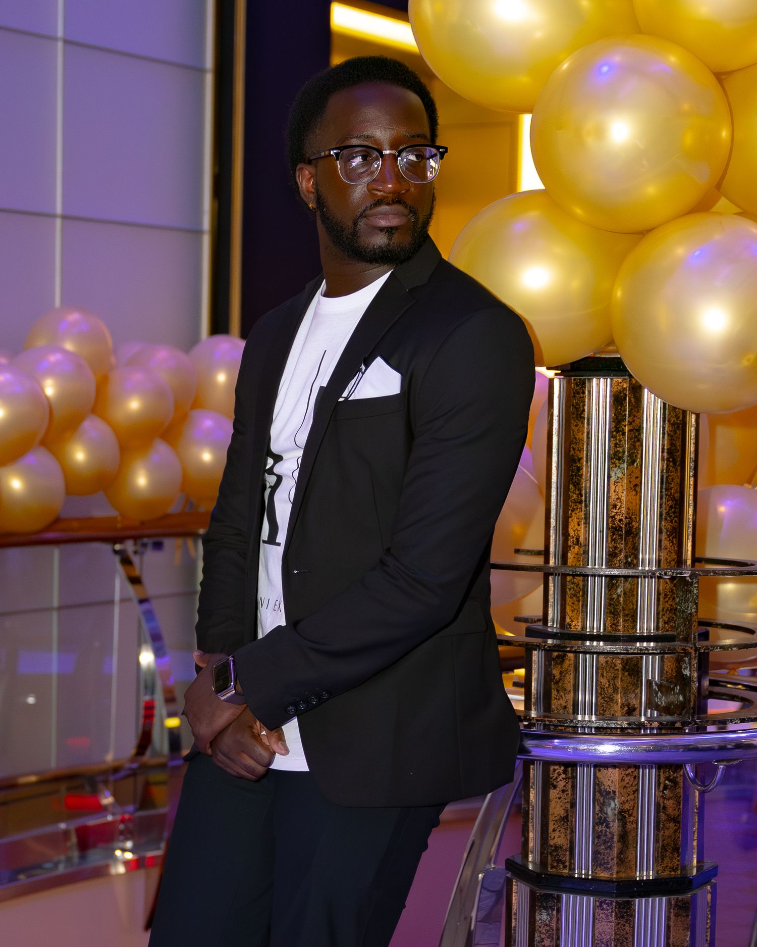 A person in a black blazer and white shirt poses at an elegant event with gold balloons in the background.