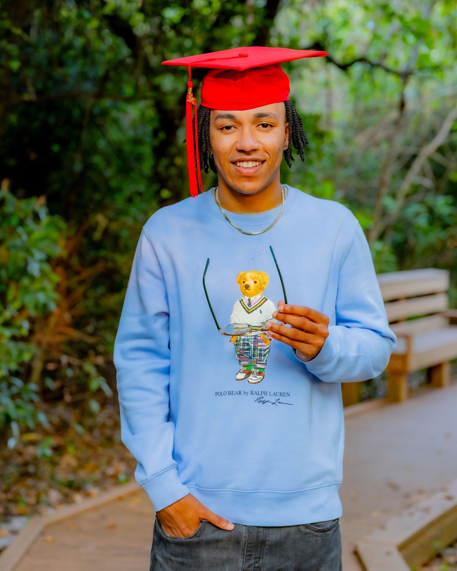 Graduate in light blue bear sweater and red graduation cap poses on outdoor pathway with greenery in background.