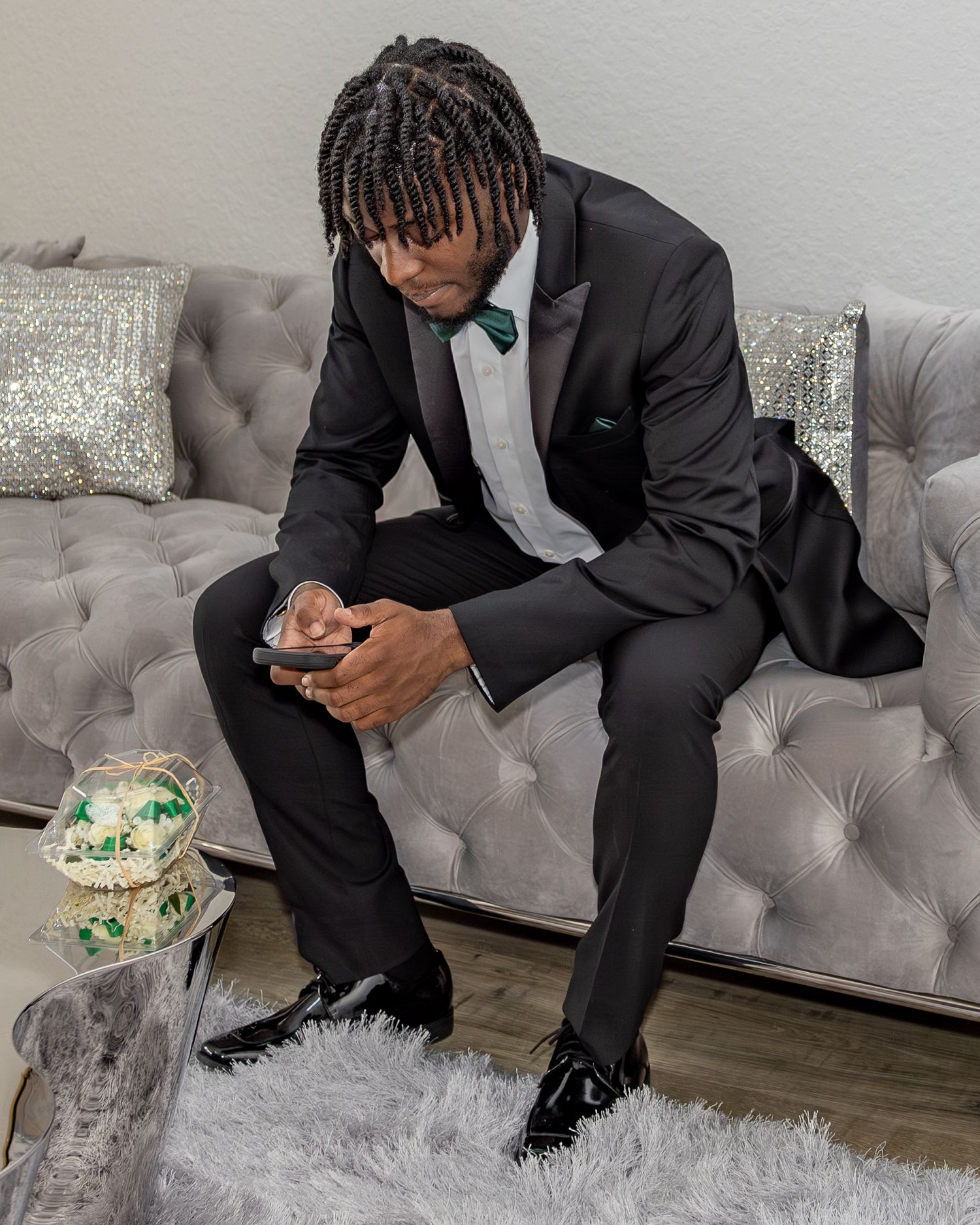 Person in a formal black suit checking phone while sitting on a gray tufted couch with decorative pillows.