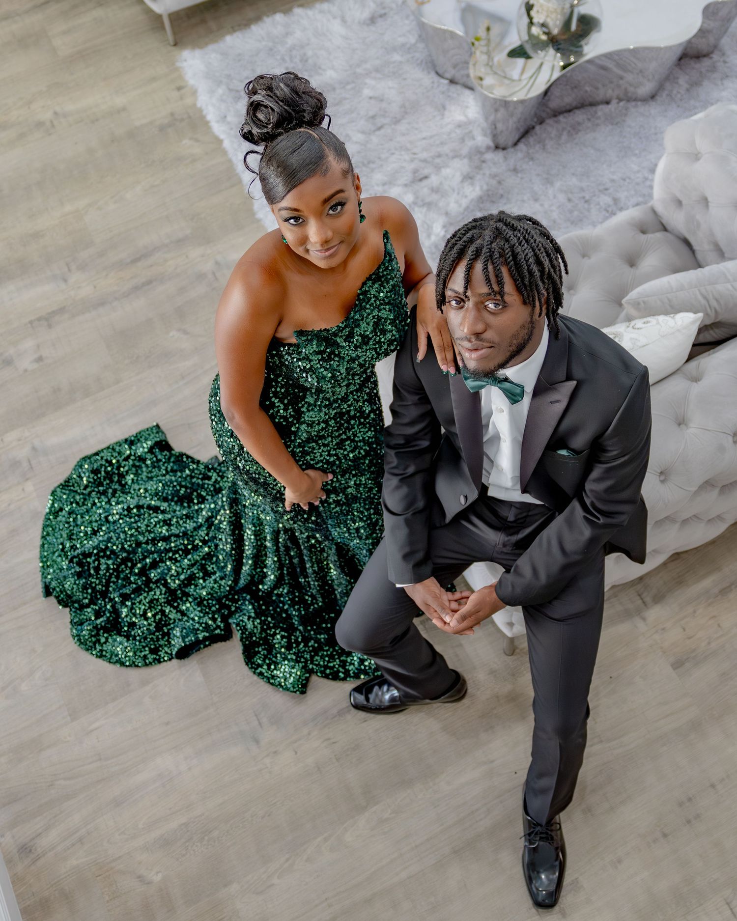 Couple in formal attire with sparkling emerald green gown and classic black tuxedo pose on light wood flooring.