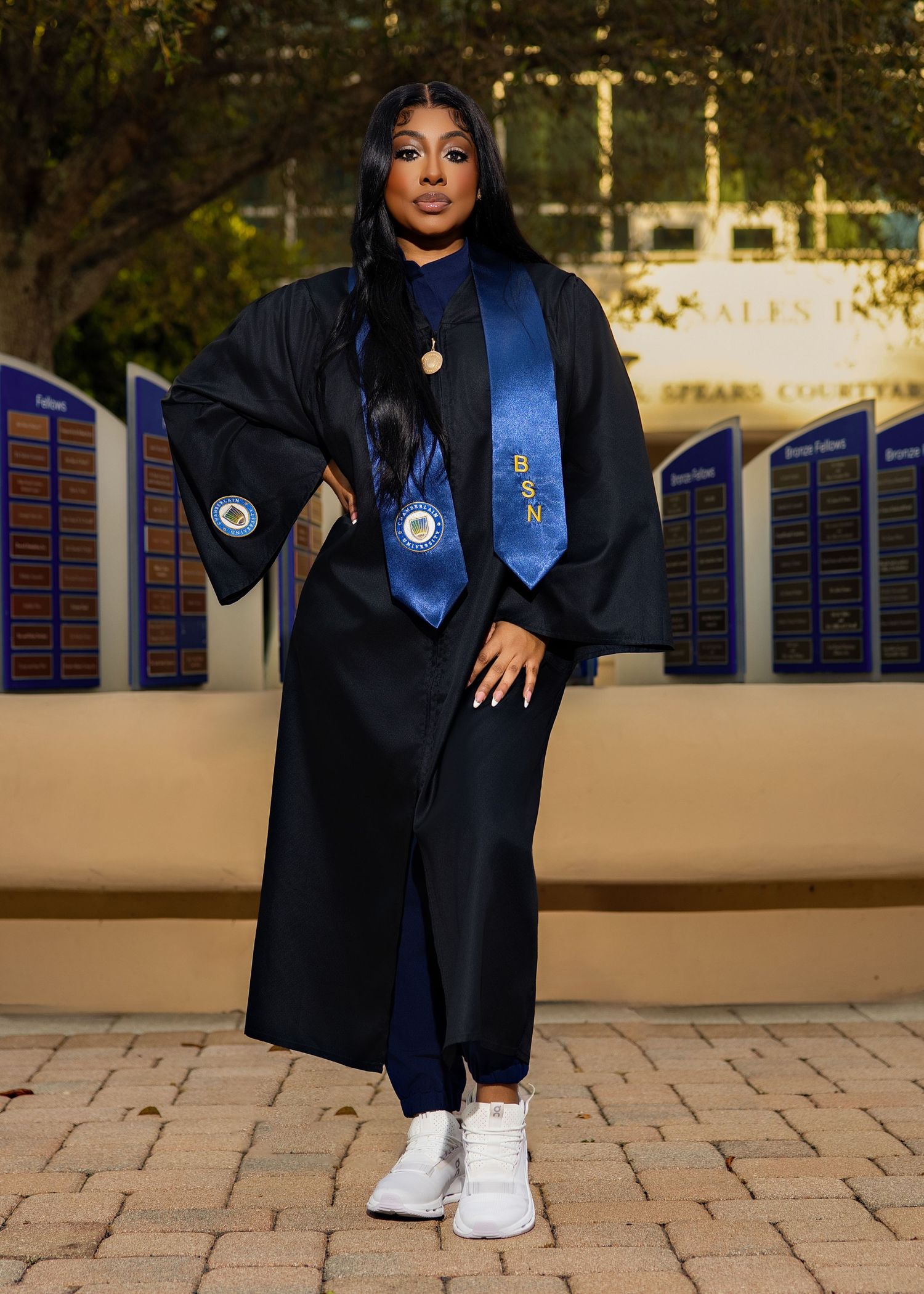 Graduate in black cap and gown with blue tie and stole poses outdoors in white sneakers for graduation photo.