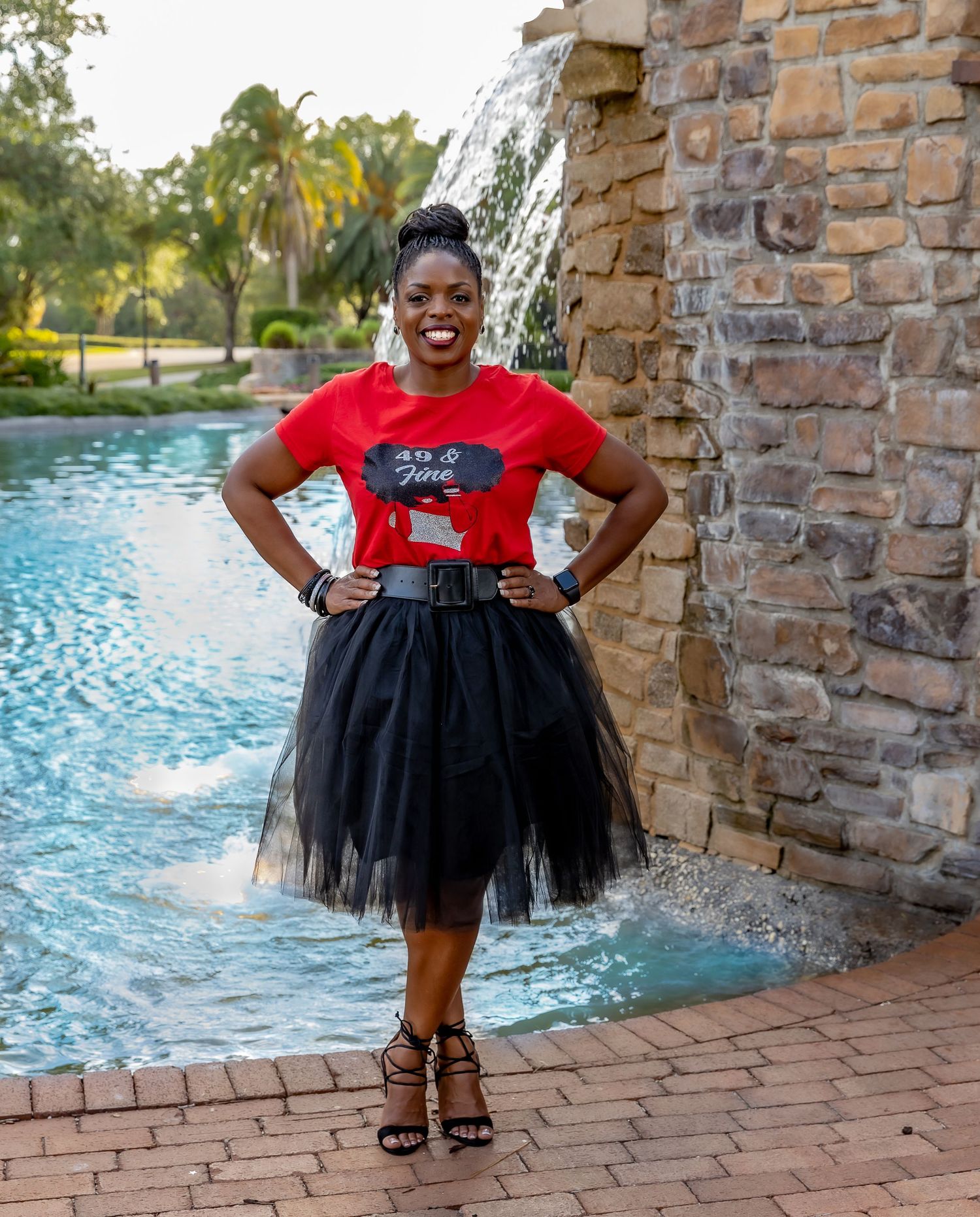 Someone poses by a stone fountain wearing a red t-shirt with black tulle skirt and strappy heels on brick pavers.
