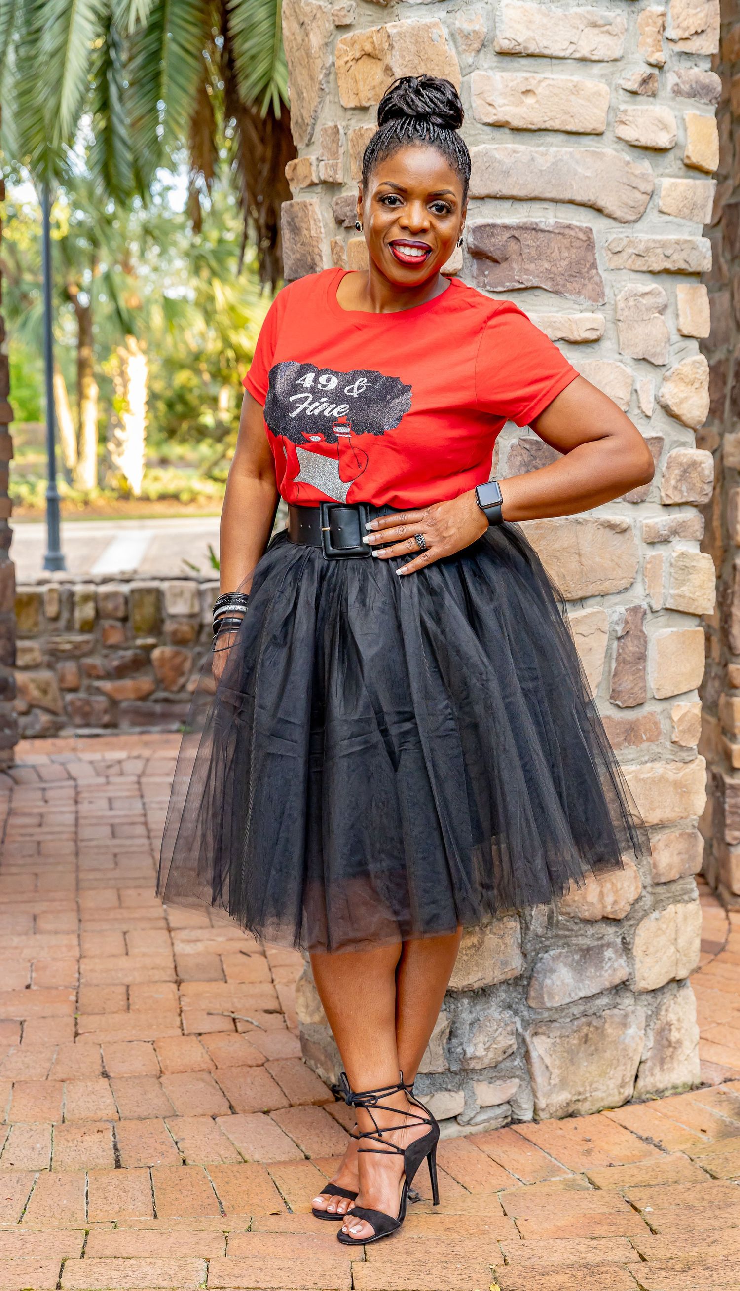 Fashion model wearing a red t-shirt with black tulle skirt poses against stone wall on brick patio.
