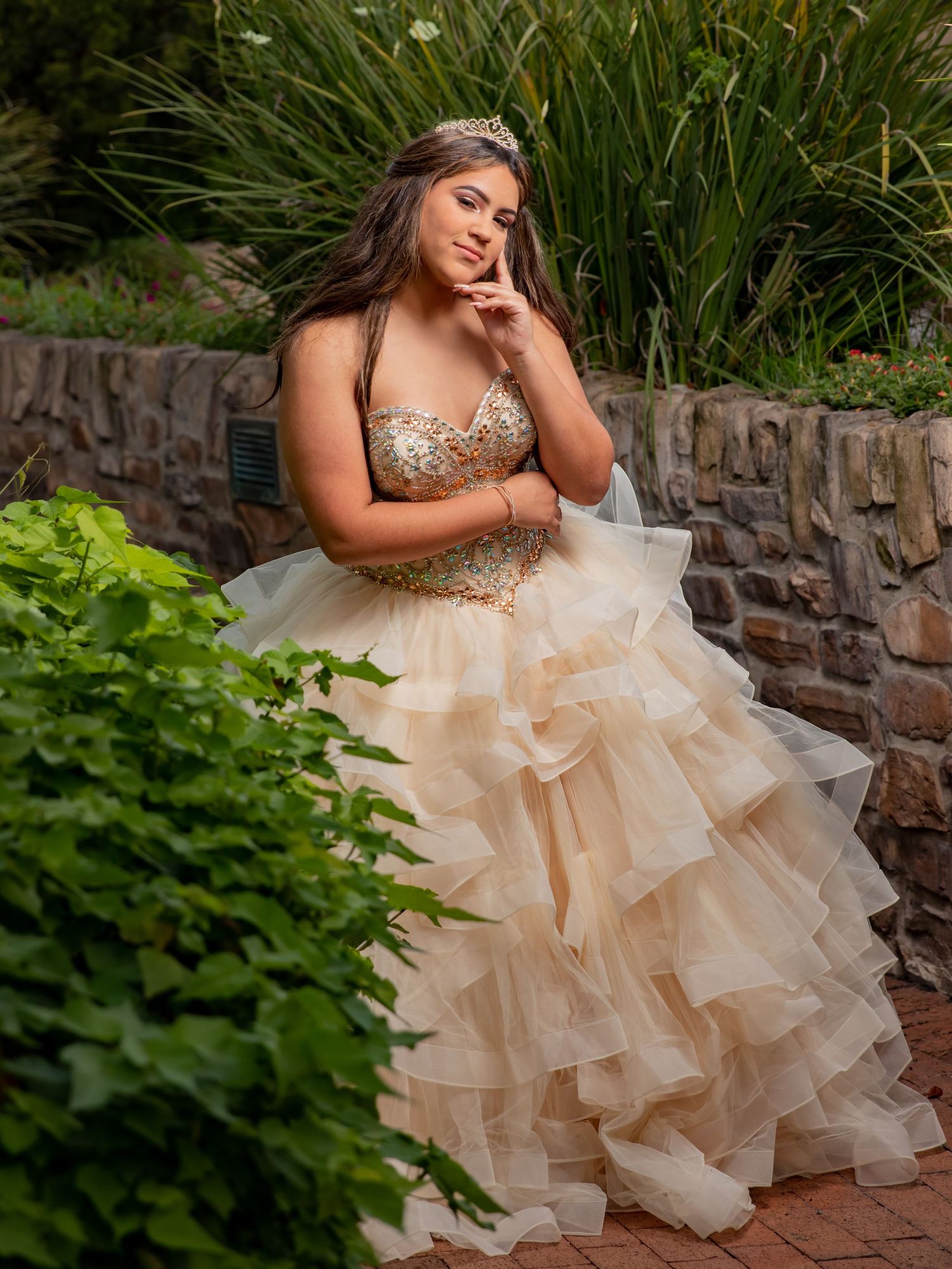 A ruffled champagne ballgown with sequined bodice photographed against stone wall and tropical foliage backdrop.