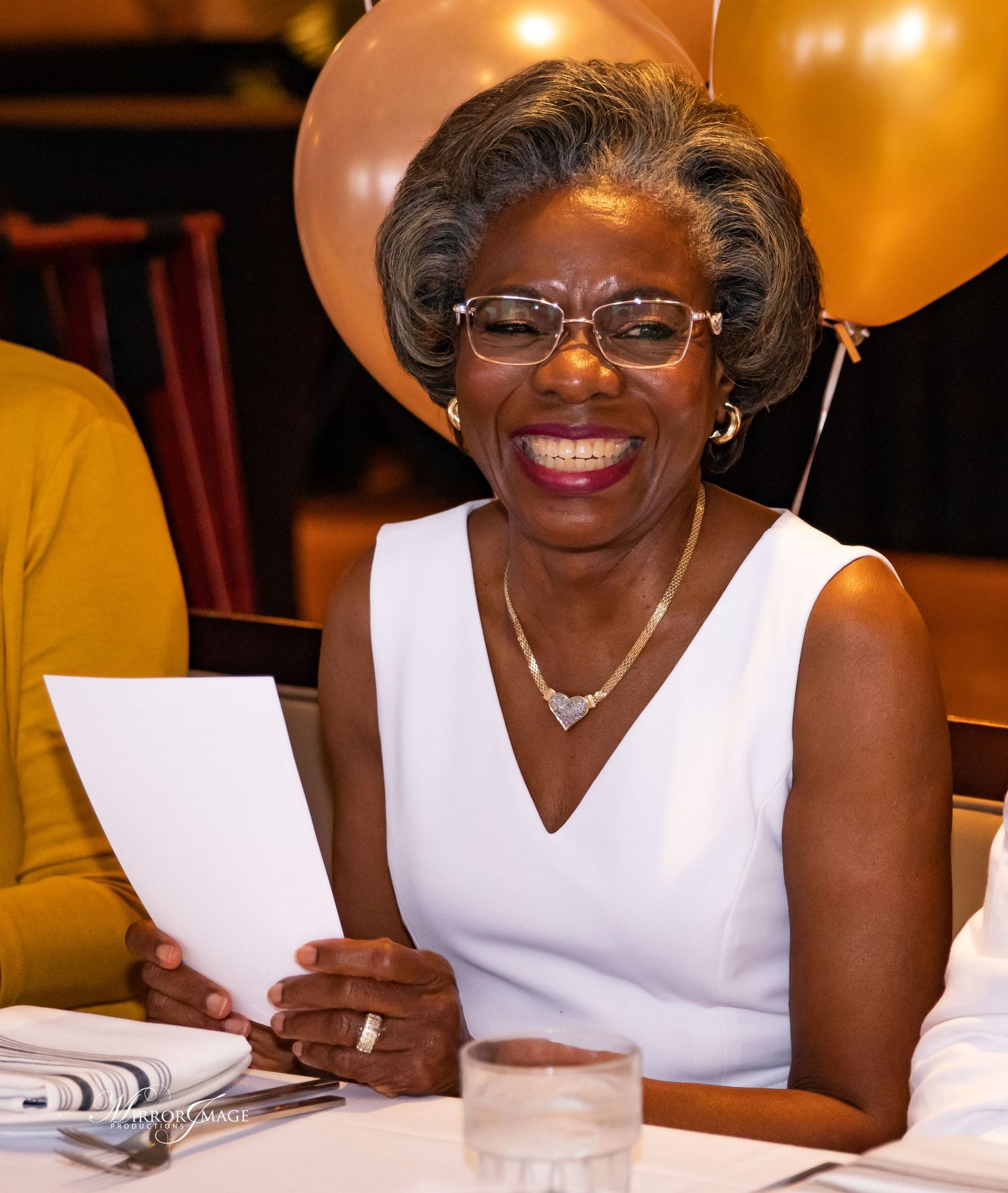 Elegant woman in white dress and glasses smiles while holding papers during a celebration with balloons in background.