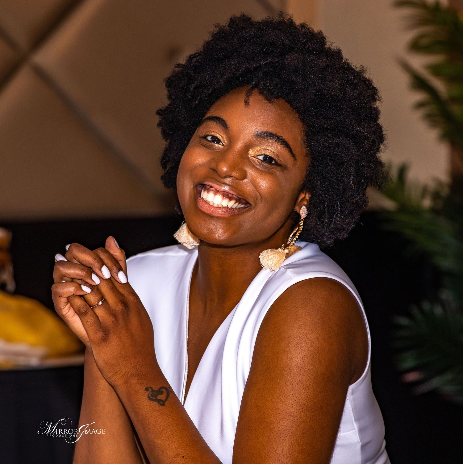 A person with natural curly black hair wearing a white sleeveless top smiles warmly while sitting indoors.