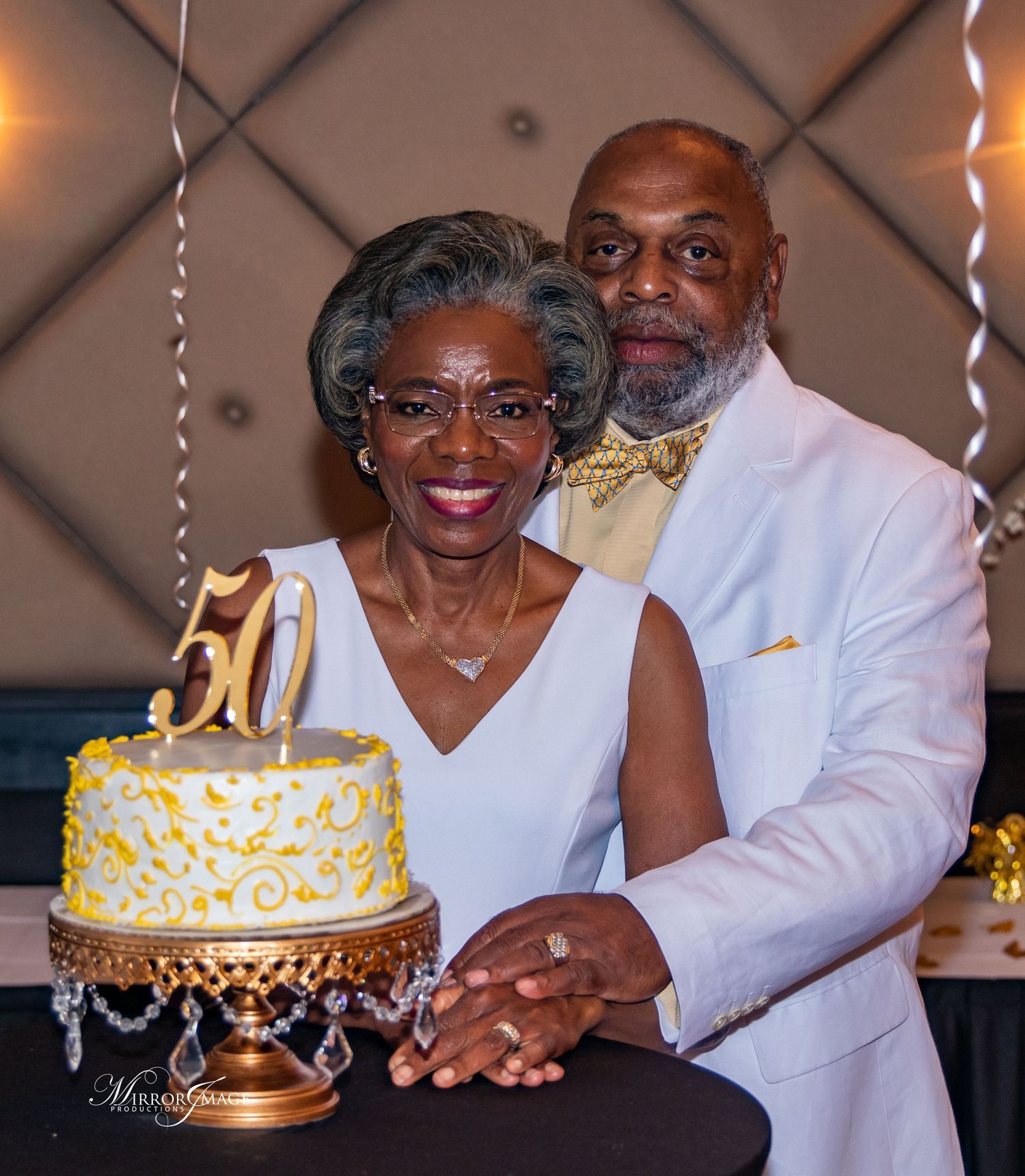 Elegant couple celebrates 50th wedding anniversary with a beautiful gold and white cake on a decorative gold stand.