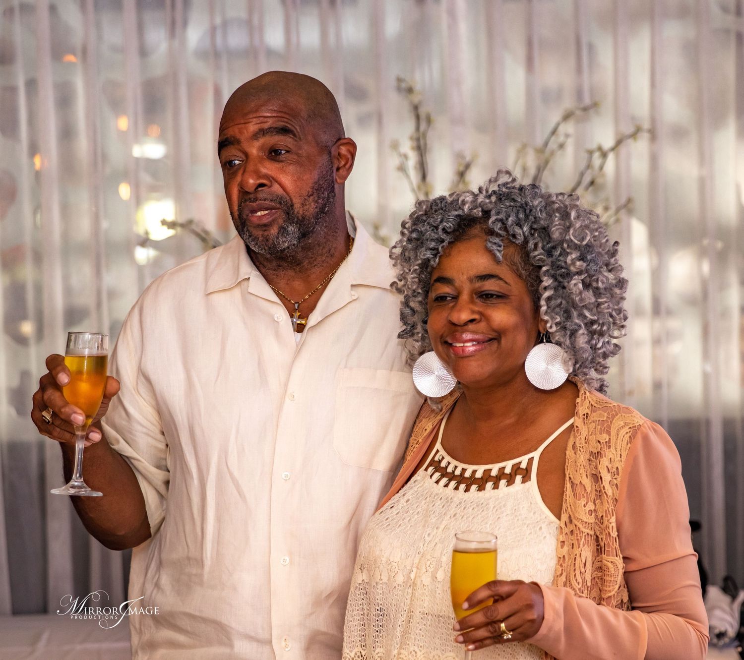 A happy couple in formal attire smiles while holding cocktails at an elegant celebration with a sparkly backdrop.