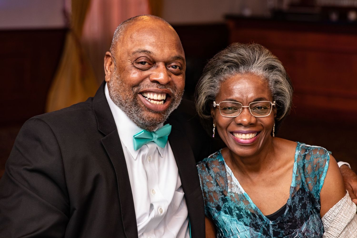 Two elegantly dressed people at a formal event smile warmly while wearing a bow tie and teal evening dress.