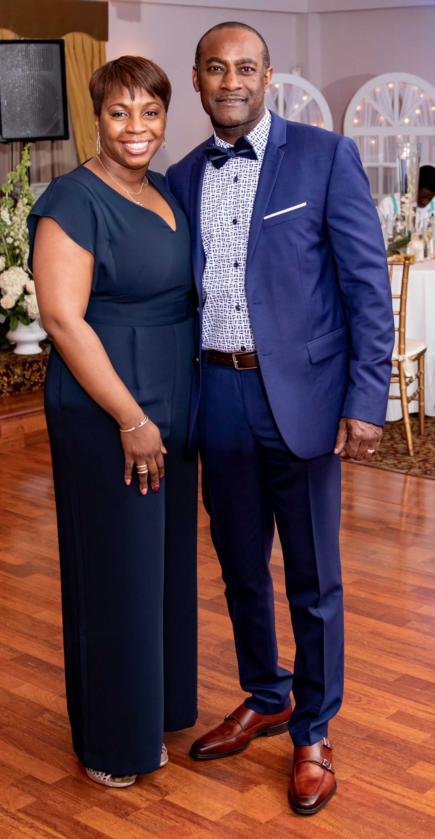 A couple in formal attire poses together at an event, wearing complementary navy blue outfits on a hardwood floor.