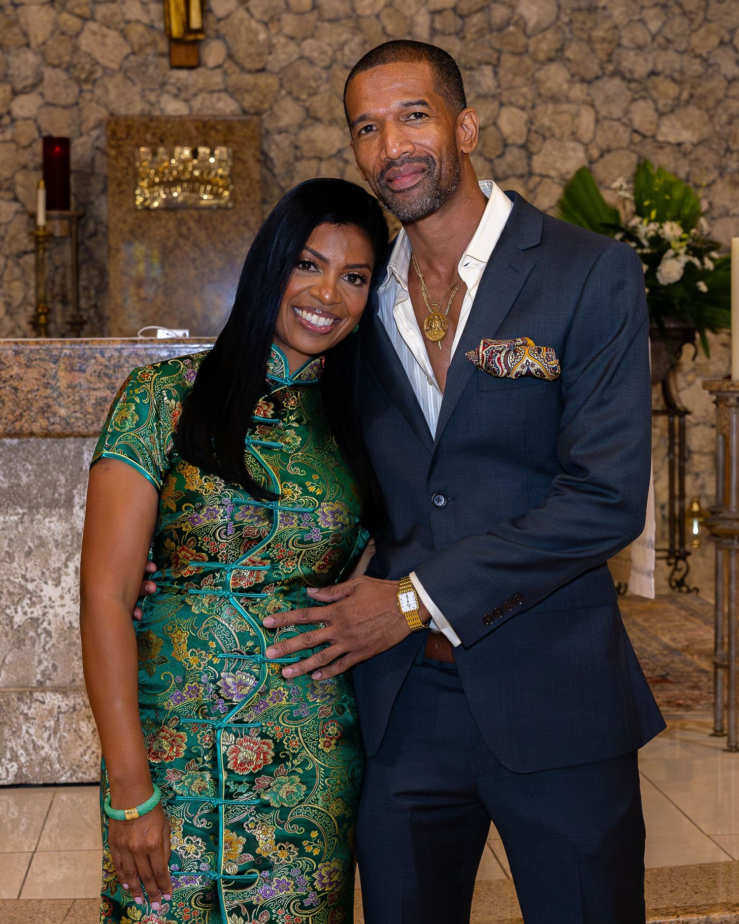 A couple in formal attire, with one wearing a navy suit and the other in a green patterned dress, pose together at an event.
