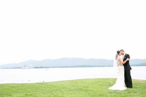 A couple in wedding attire embraces on a grassy lawn overlooking a serene lake and mountains.