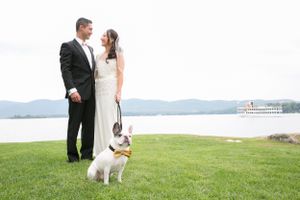 A couple in wedding attire stands on a grassy lawn by a lake with their small white dog on a leash.