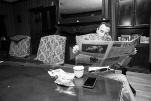 A person sits at a dining table reading a newspaper with a coffee cup and smartphone nearby in black and white.