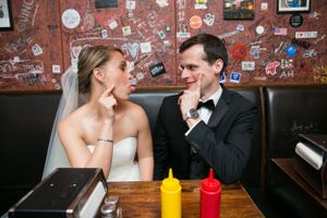 Newlyweds share an intimate moment at a casual restaurant booth with red and yellow condiment bottles on wooden table.