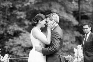 A black and white photo captures a tender moment between newlyweds sharing a kiss during an outdoor wedding ceremony.