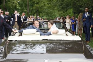 A couple wave from inside a vintage convertible car while surrounded by wedding guests on a tree-lined path.