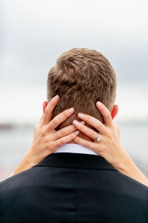 Back view of a wedding couple with hands embracing the back of the groom's head showing an engagement ring.