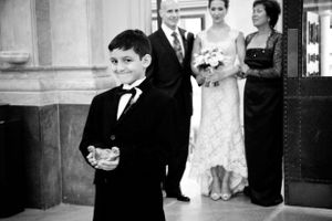 Black and white photo of a young ring bearer in a tuxedo at a wedding ceremony with the bridal party behind him.