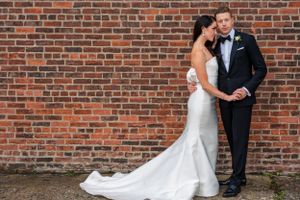 A couple in wedding attire stands against a rustic brick wall, with the bride's white gown flowing elegantly behind her.