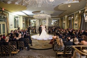 A grand ballroom wedding ceremony with crystal chandeliers and guests seated in black chairs facing a circular platform.