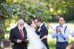 A joyful wedding ceremony takes place outdoors under green foliage on a sunny summer day.