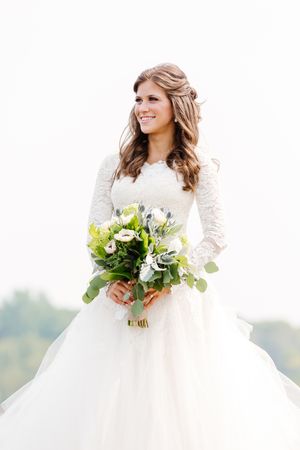 Bride in long-sleeved lace wedding gown holds green and white bouquet while smiling outdoors.