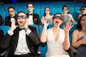 Wedding party wearing 3D glasses reacts with excitement while watching a movie in theater seats.