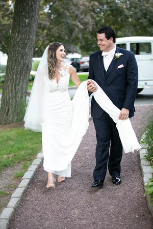 A newlywed couple walks down a tree-lined path while laughing and holding their wedding attire.